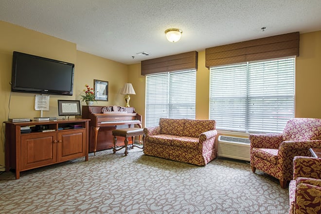 A cozy living room with a piano, sofa, armchair, TV, and large windows.