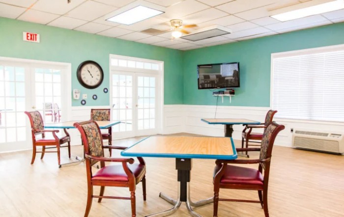 A bright dining area with several tables and chairs, featuring a wall-mounted television and a clock.