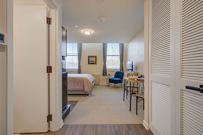 A cozy bedroom with a neatly made bed, a blue chair, and large windows letting in natural light.