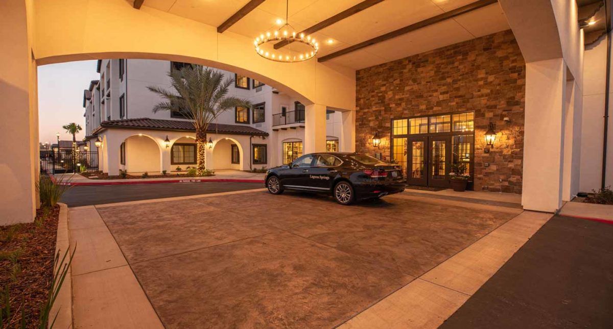 A luxury car parked at the entrance of an upscale hotel, illuminated by elegant evening lighting and a grand chandelier.