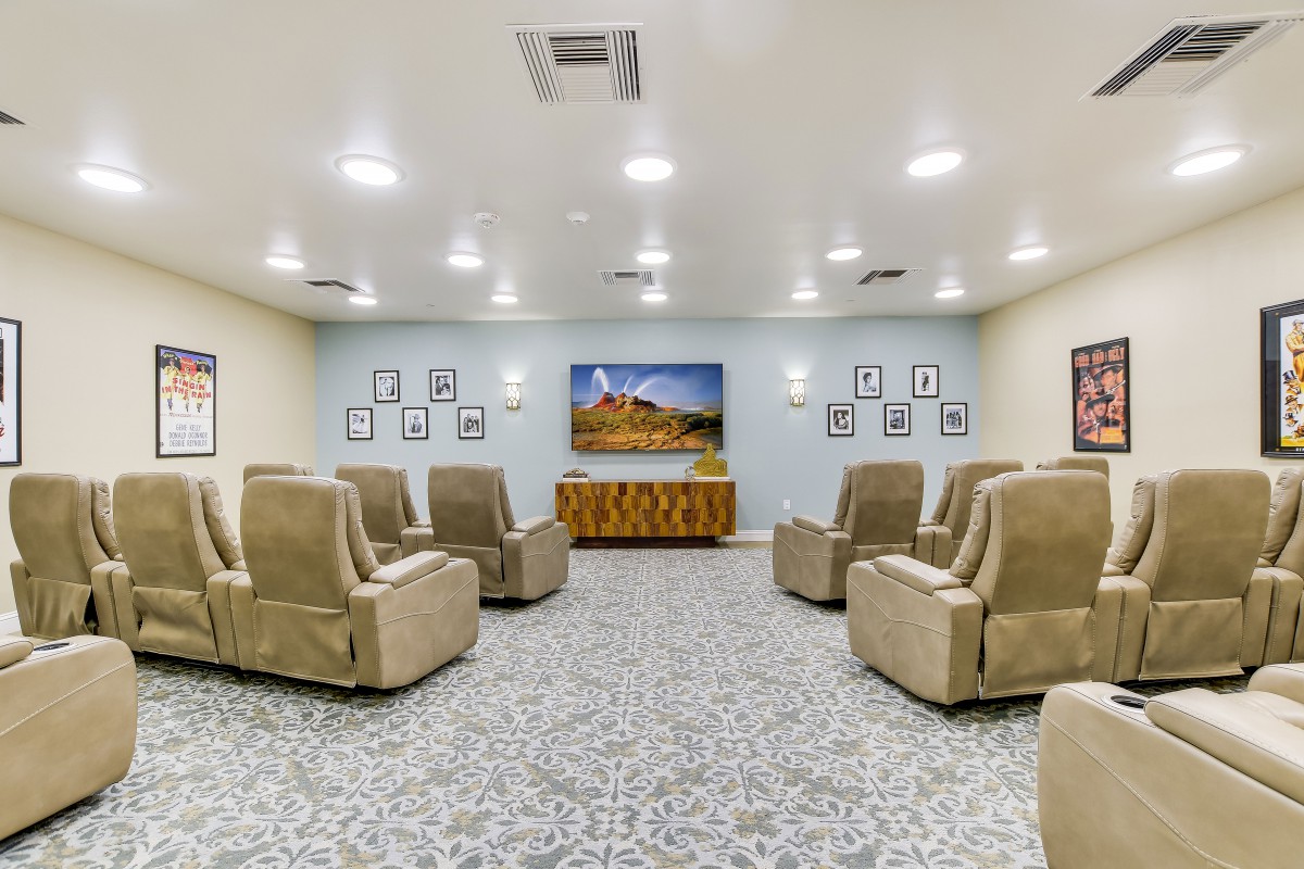 A cozy theater room with beige recliner chairs facing a large television on a patterned carpet floor, surrounded by soft lighting.