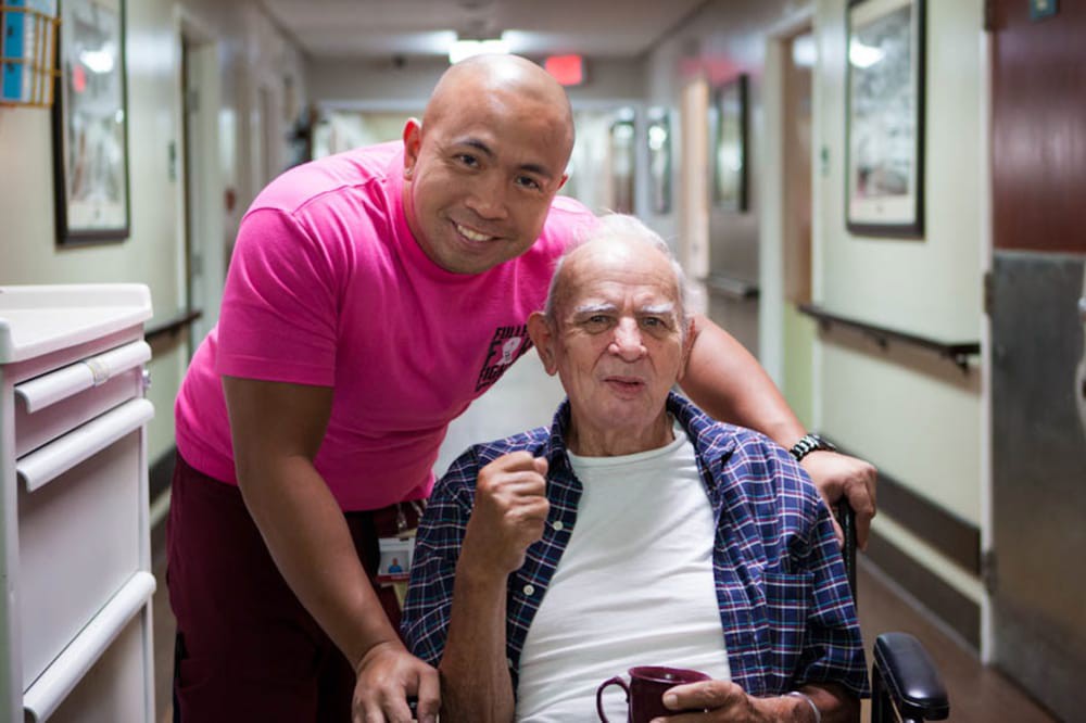 A caregiver poses with an elderly man in a wheelchair inside a nursing home.