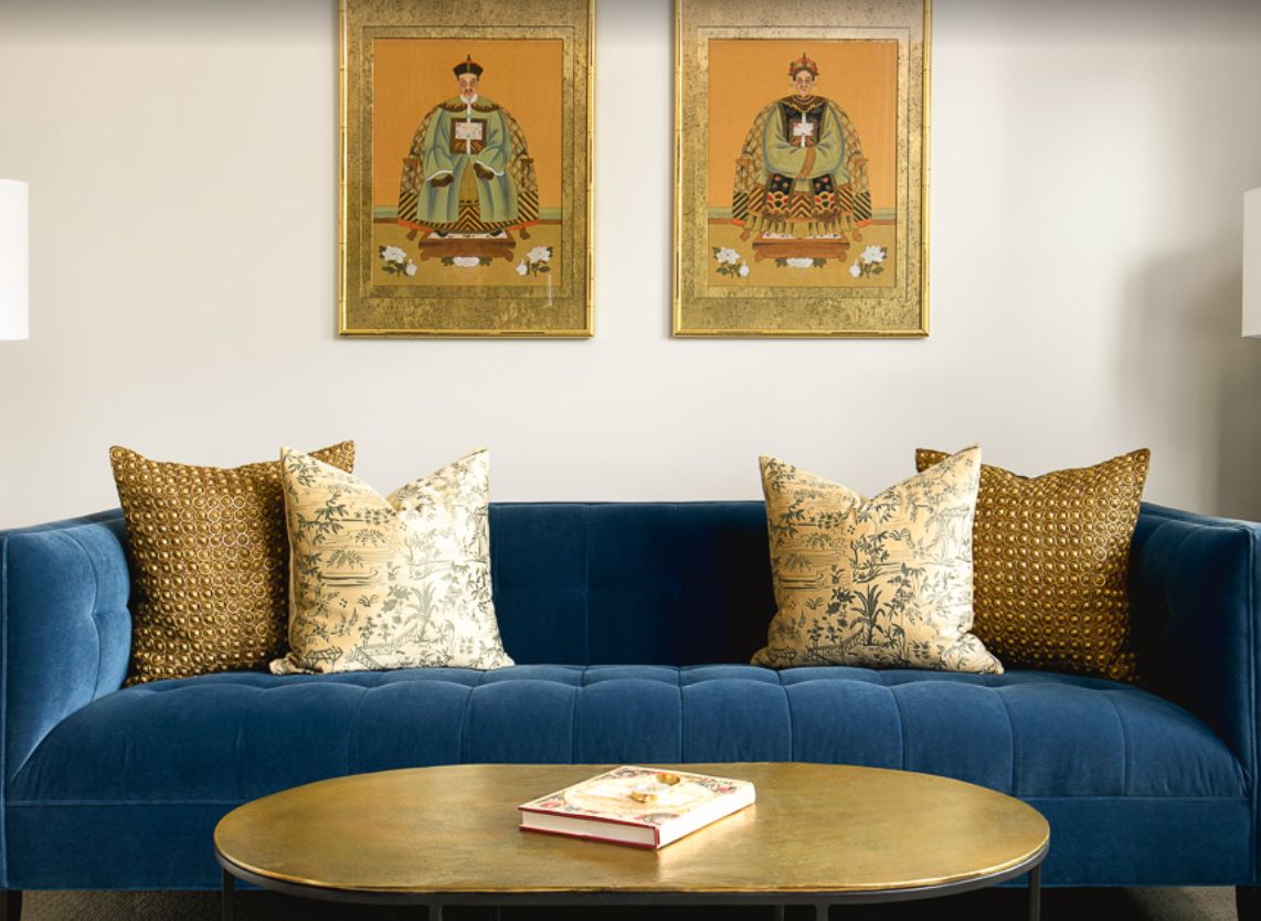 A stylish living room featuring a blue sofa with patterned pillows, two framed artworks on the wall, and a round coffee table with a book.