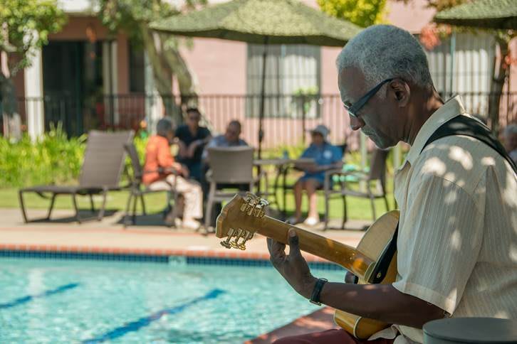 A man plays guitar by the poolside while others relax and enjoy the sunny day.