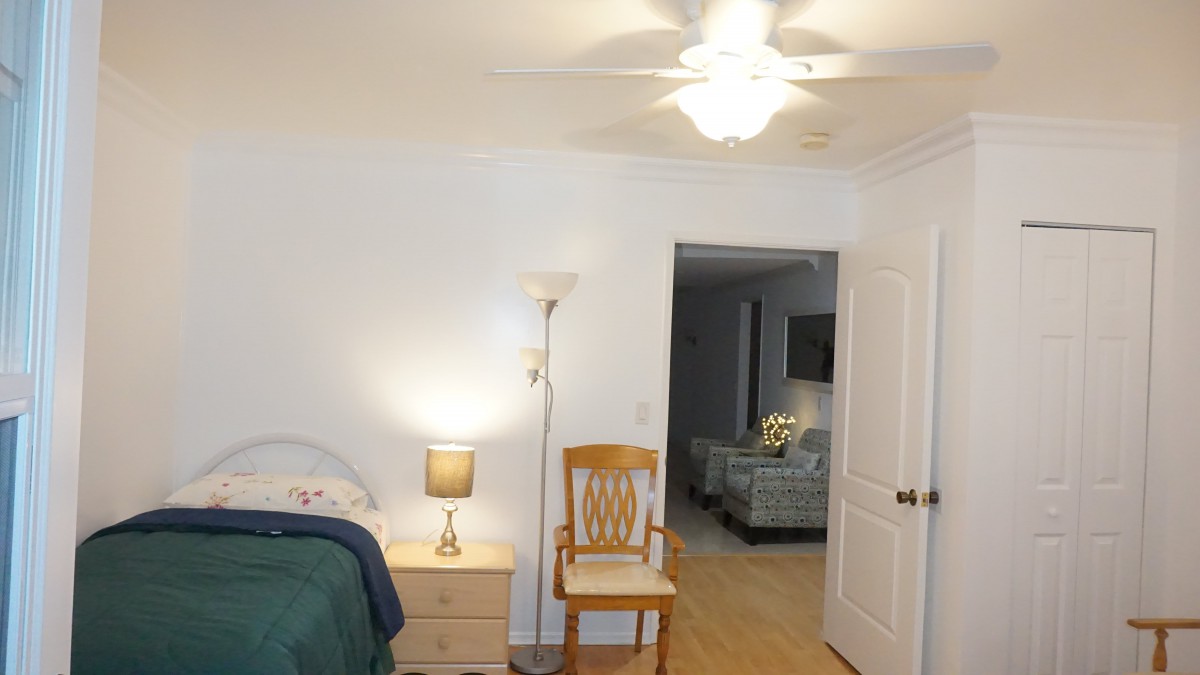 A cozy bedroom with a wooden chair, bedside table, and lamps, leading into a living area.