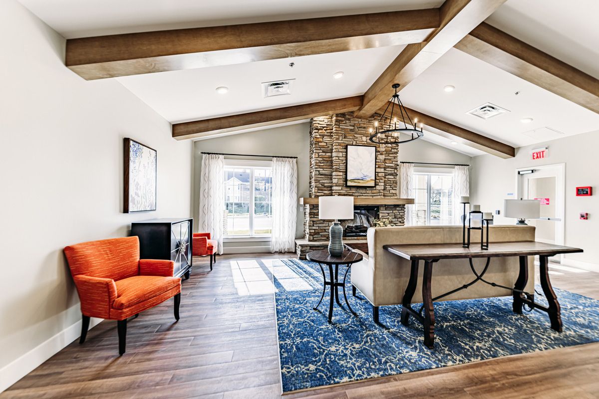 A modern living room with vaulted ceilings, wood beams, and a stone fireplace, featuring orange chairs and a blue patterned rug.