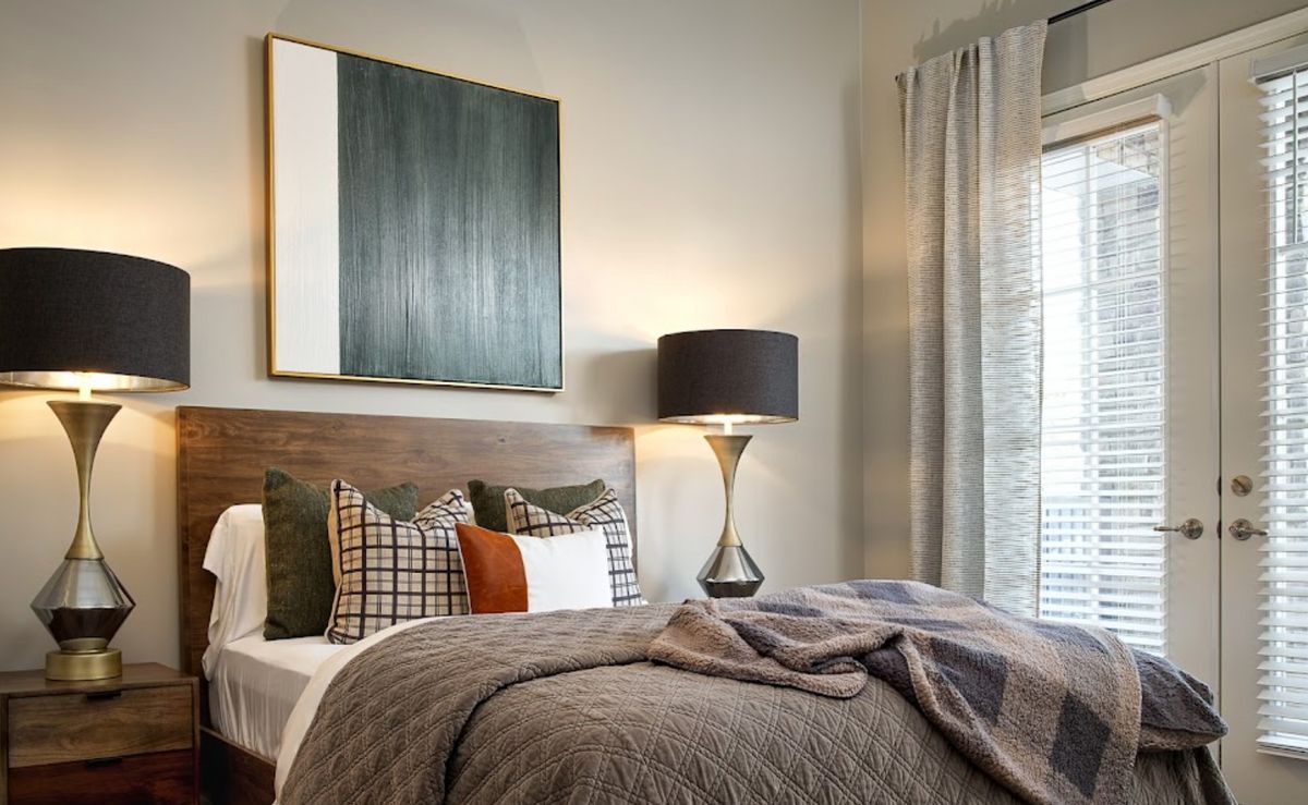A modern bedroom with stylish decor, featuring a large artwork above the bed, a sleek lamp on the side table, and cozy pillows and comforter.