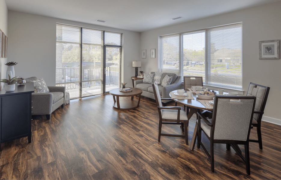 A modern living room with hardwood floors and ample natural light, featuring a cozy seating area and a dining space by large windows.