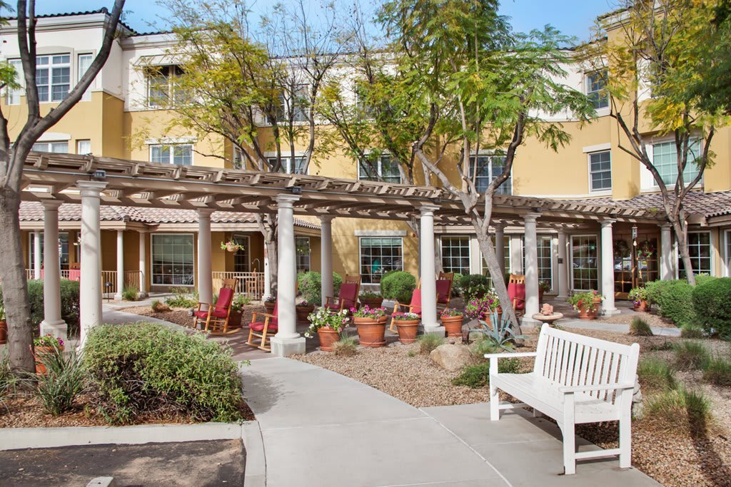 A peaceful outdoor garden area with benches, chairs, and surrounding greenery in front of a yellow building.