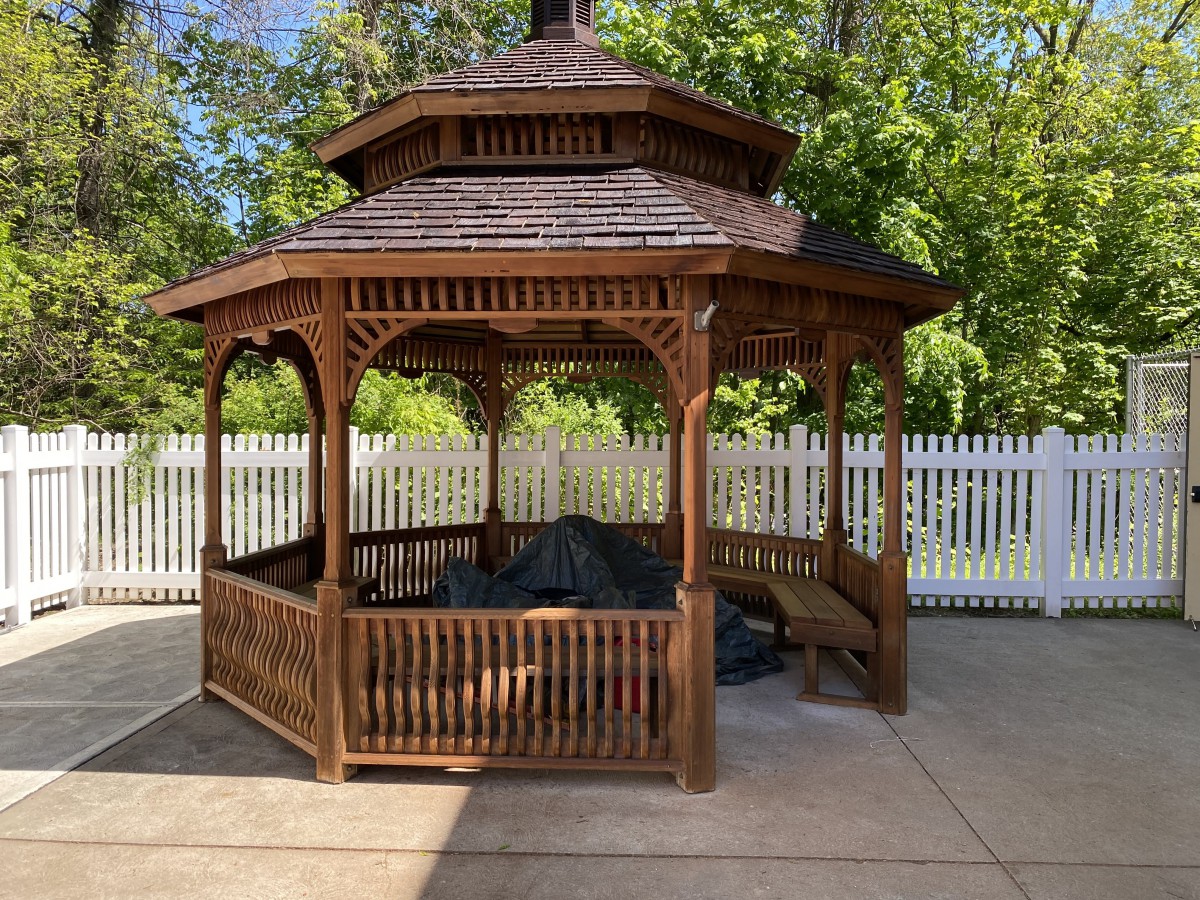 A wooden gazebo surrounded by a white picket fence and greenery.