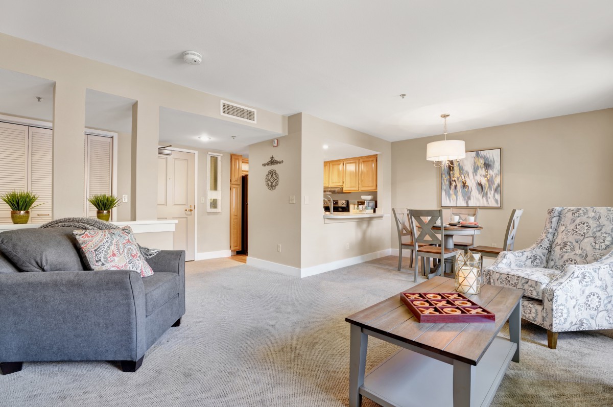 A cozy living room with a sofa, table, and dining area, featuring warm decor and ample lighting.
