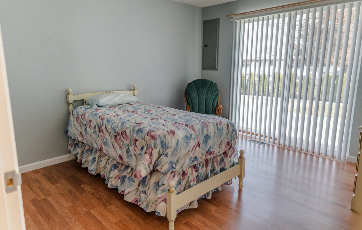 A simple bedroom with a single bed covered in floral bedding, a green chair, and large window blinds letting in daylight.