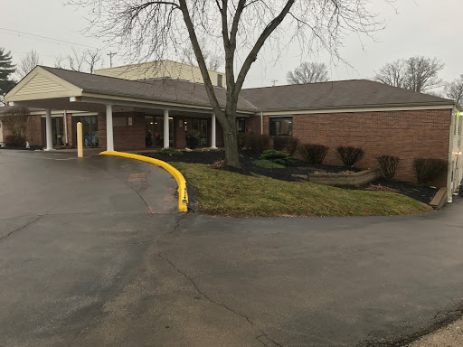 A view of a low-rise brick building with a parking area, featuring landscaping and a prominent tree.