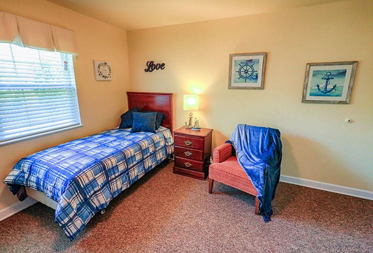 A cozy bedroom with a single bed and an armchair beside a small bedside table, illuminated by a lamp.