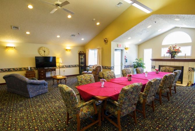 A spacious and well-lit living and dining area featuring a long table with red tablecloths, comfortable chairs, a television, indoor plants, and ample natural light.