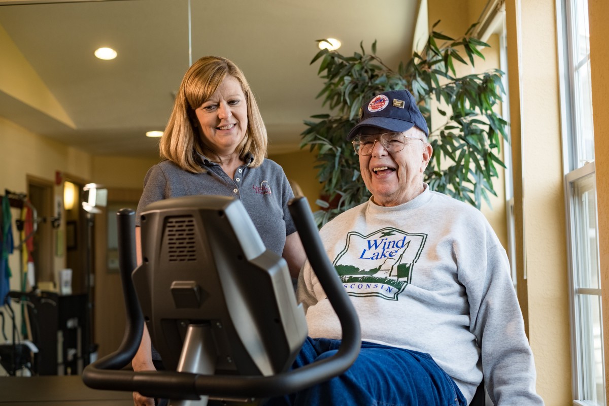 An elderly man smiles while exercising on a stationary bike, accompanied by a woman indoors.
