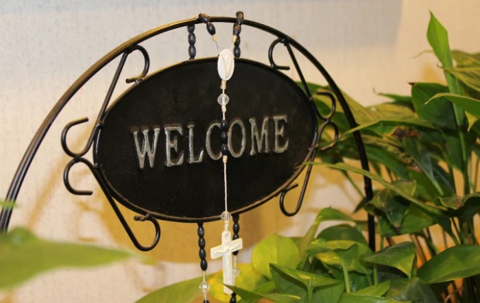 A welcome sign adorned with a rosary and cross, surrounded by indoor plants.