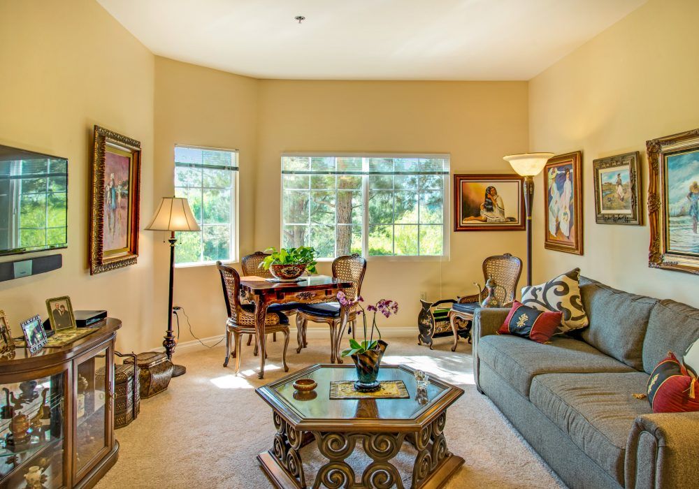 A cozy and elegantly furnished living room with large windows, featuring a sofa, coffee table, dining area, and decorative artworks.