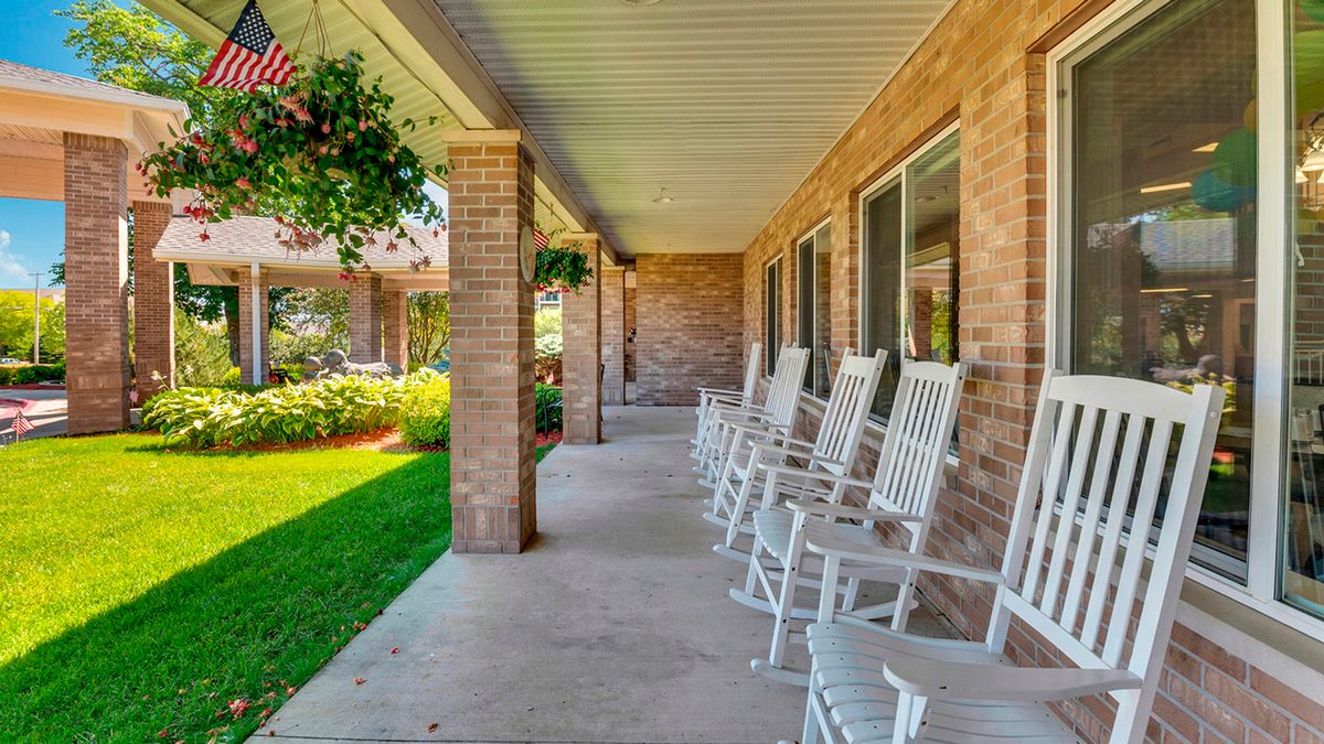 A welcoming porch with white rocking chairs and hanging plants, attached to a brick building.