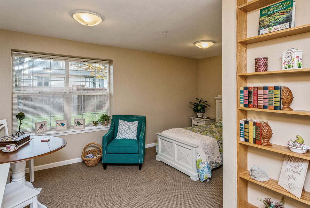 A cozy bedroom with a teal armchair, a bed, and a bookshelf under bright lighting.