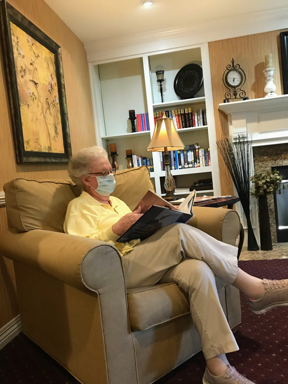 A person wearing a mask is sitting in a chair reading a book in a cozy living room.