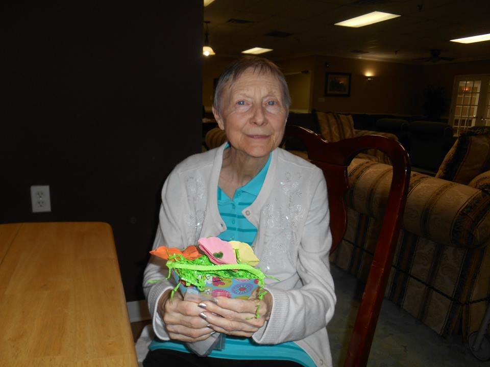 An elderly woman smiling while holding a colorful craft indoors.