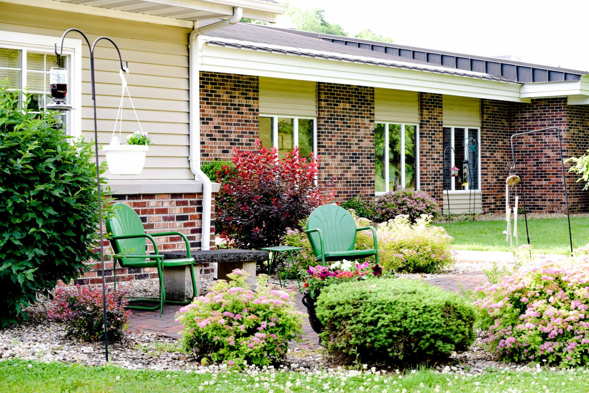A cozy garden patio with green chairs nestled among lush shrubs and plants in front of a brick house.