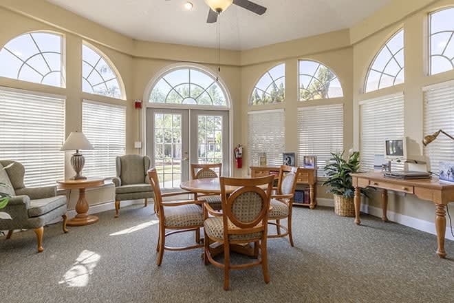 A well-lit dining area with a circular table and chairs, featuring large windows and indoor plants.