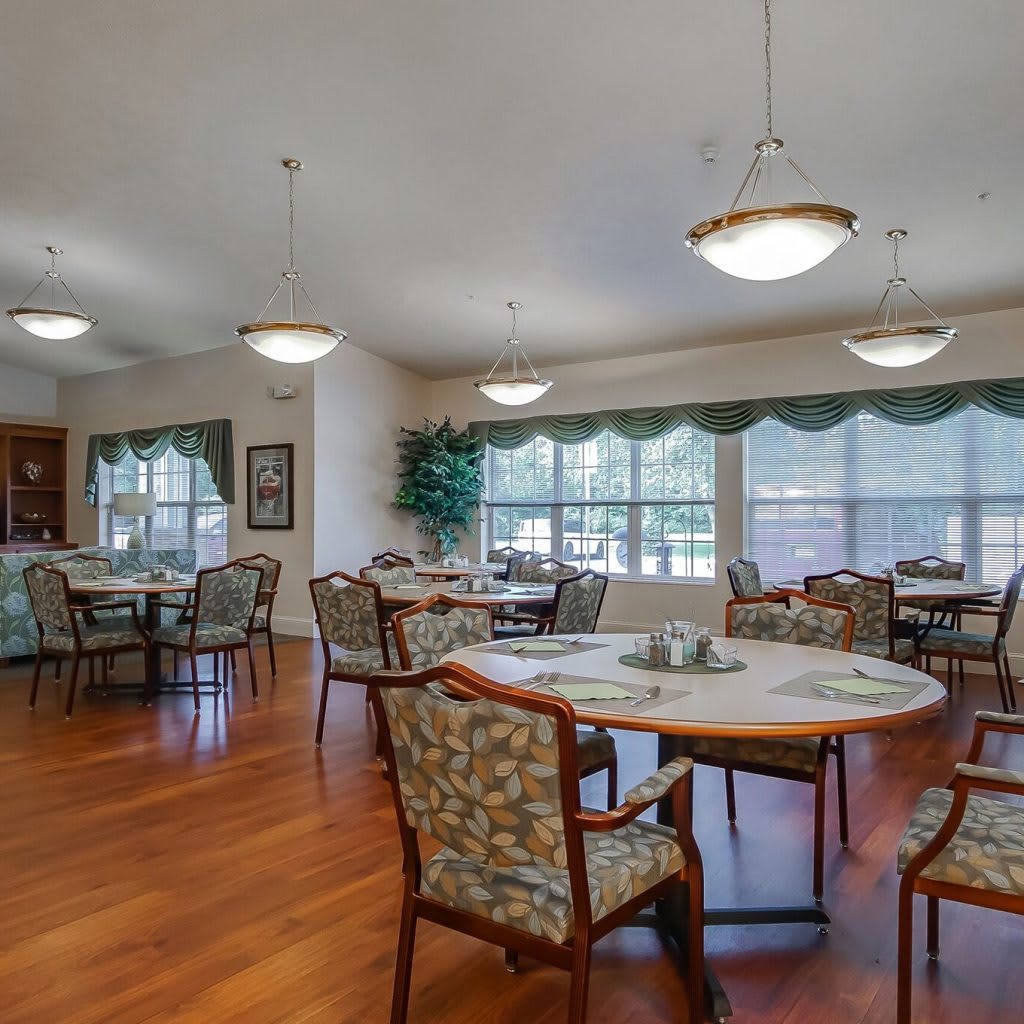 A spacious dining room with round tables and chairs, large windows, and wooden flooring.