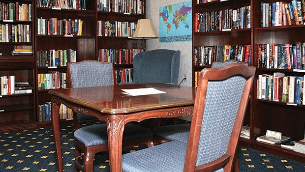 A cozy library room featuring a wooden table surrounded by chairs, with bookshelves and a world map on the wall.