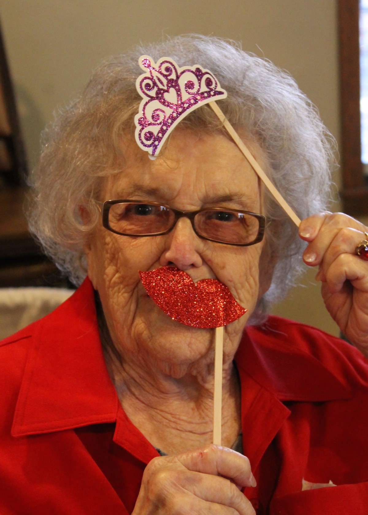 An elderly person having fun with party props, including a fake tiara and oversized red lips.