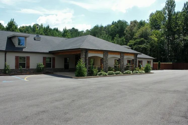 A single-story building with a parking lot in front, surrounded by trees and under a cloudy sky.