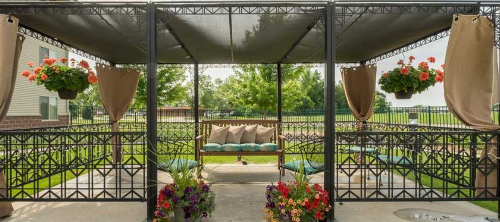 A serene outdoor gazebo with a swing, adorned with pillows and surrounded by vibrant flowers in a garden setting.