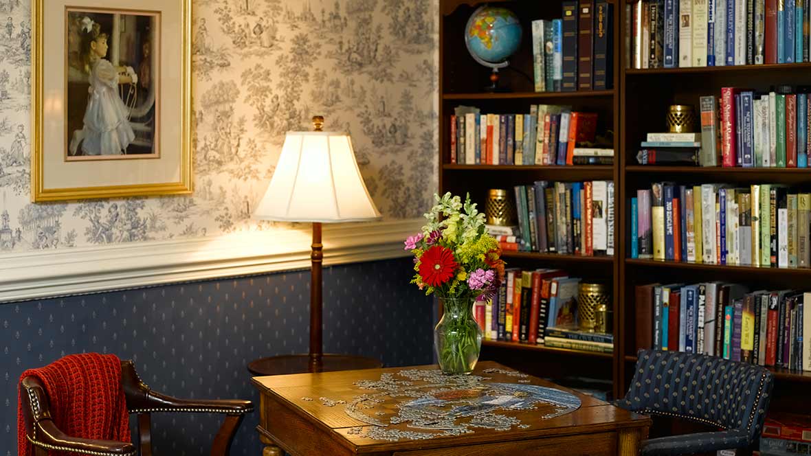 A cozy reading nook with a wooden table, adorned with a floral arrangement, next to a well-stocked bookshelf and lamp.