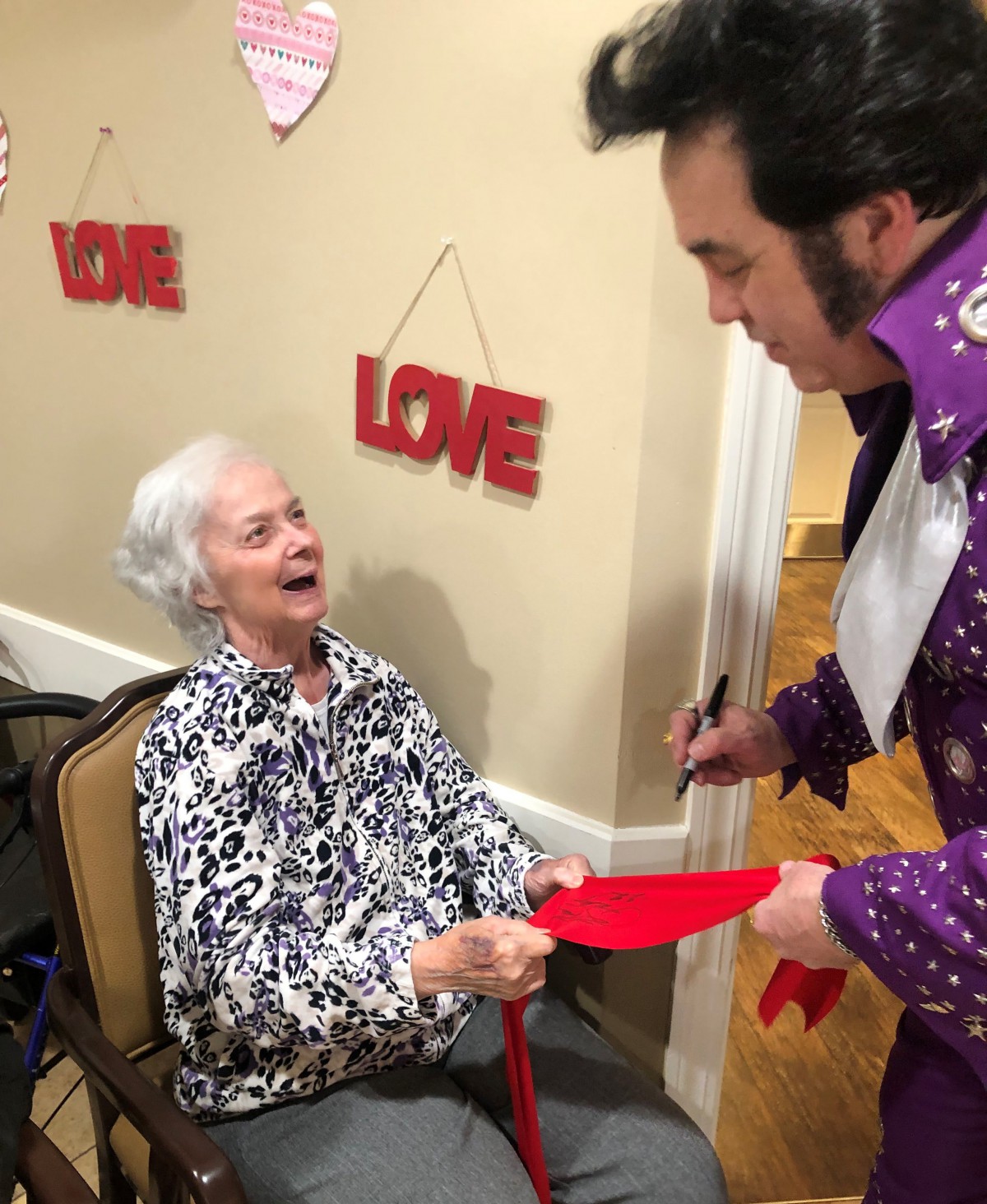 An elderly woman joyfully interacts with an entertainer wearing a colorful costume, set against a backdrop of love-themed decorations.