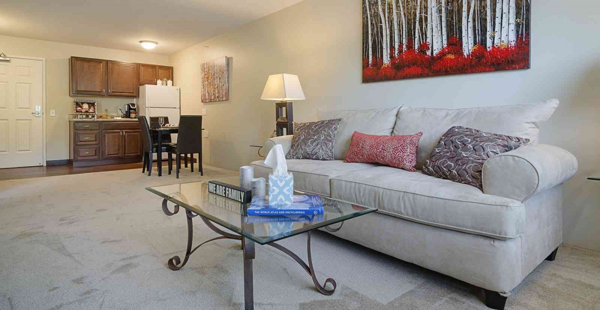 A cozy living room with a sofa, glass coffee table, and dining area in the background.