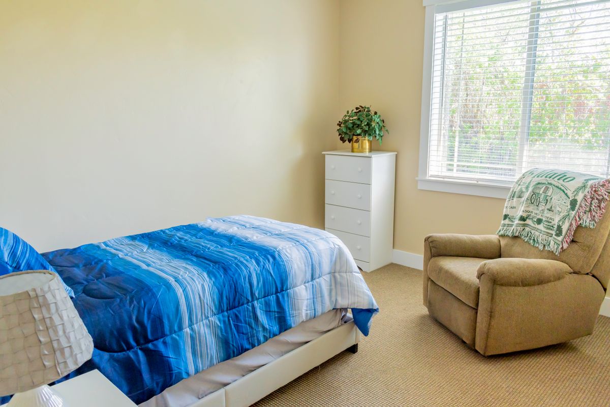 A simple bedroom featuring a single bed with blue bedding, an armchair, and a dresser near a window with natural light.