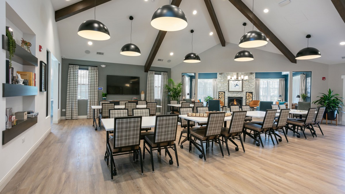 A spacious and well-lit dining or meeting area with multiple tables and chairs, featuring modern lighting fixtures and wooden ceiling beams.