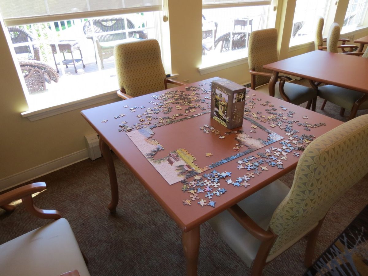 An indoor room featuring a table with a partially completed puzzle, surrounded by chairs and illuminated by natural light from the windows.