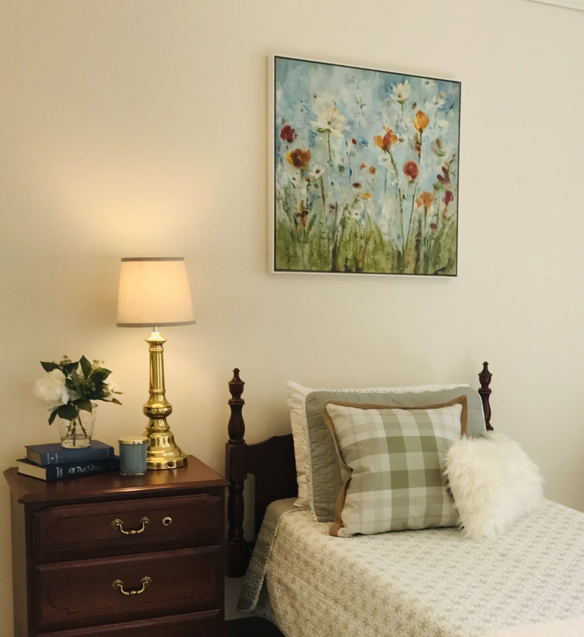 A cozy bedroom with a wooden nightstand, elegant lamp, and a floral painting above the bed.