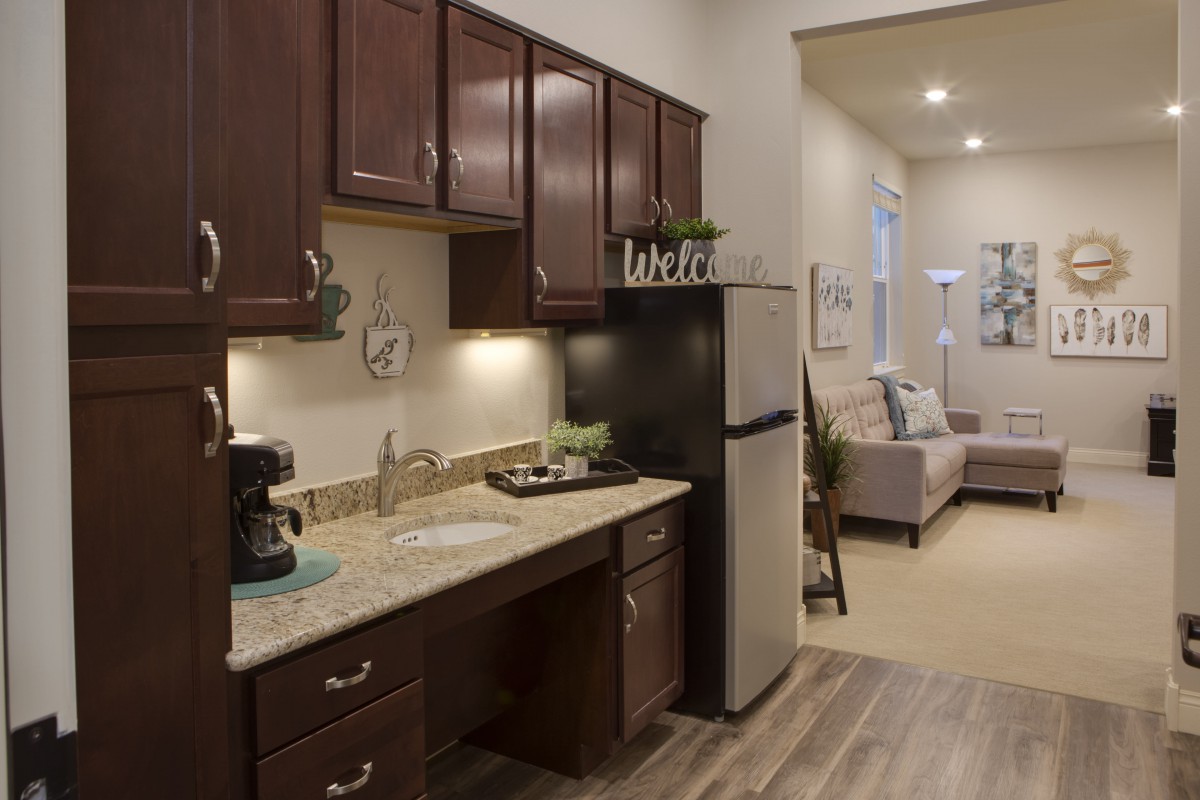 A modern apartment kitchen with wooden cabinets and a small living area in the background.