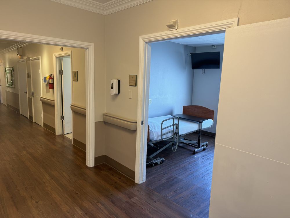 A hospital corridor with a view into a patient room containing a bed.