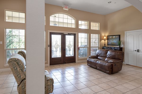A living room with a tile floor, featuring a sofa, large windows, double doors, and wall artwork.