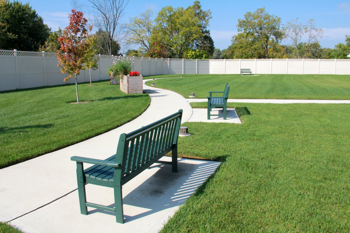 A serene park setting with green benches along a winding pathway surrounded by lush grass and trees.