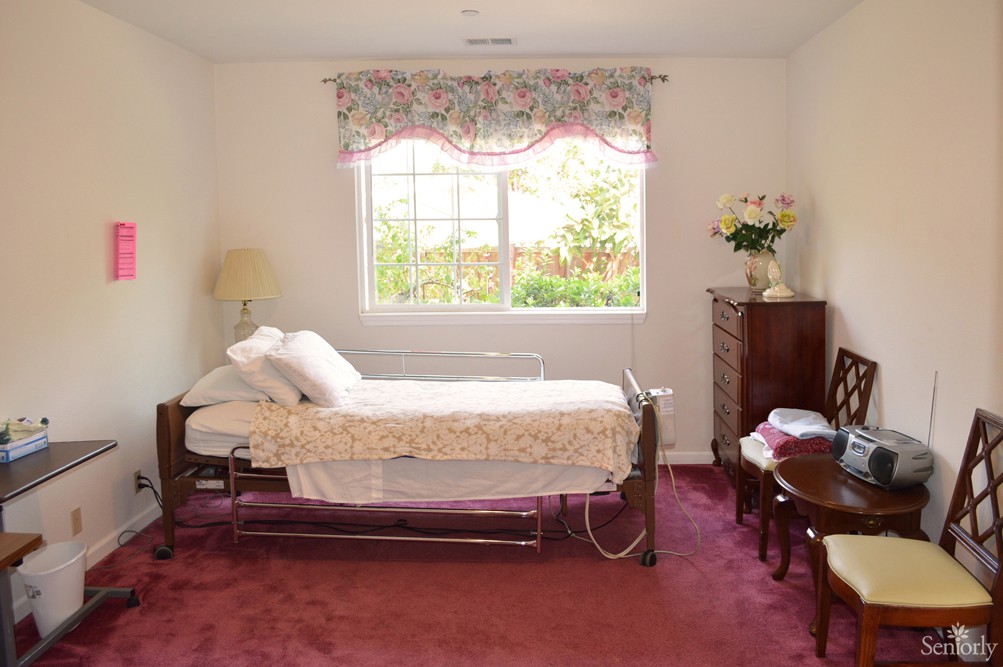 A simple bedroom with a hospital bed, decorated with floral curtains and a burgundy carpet.