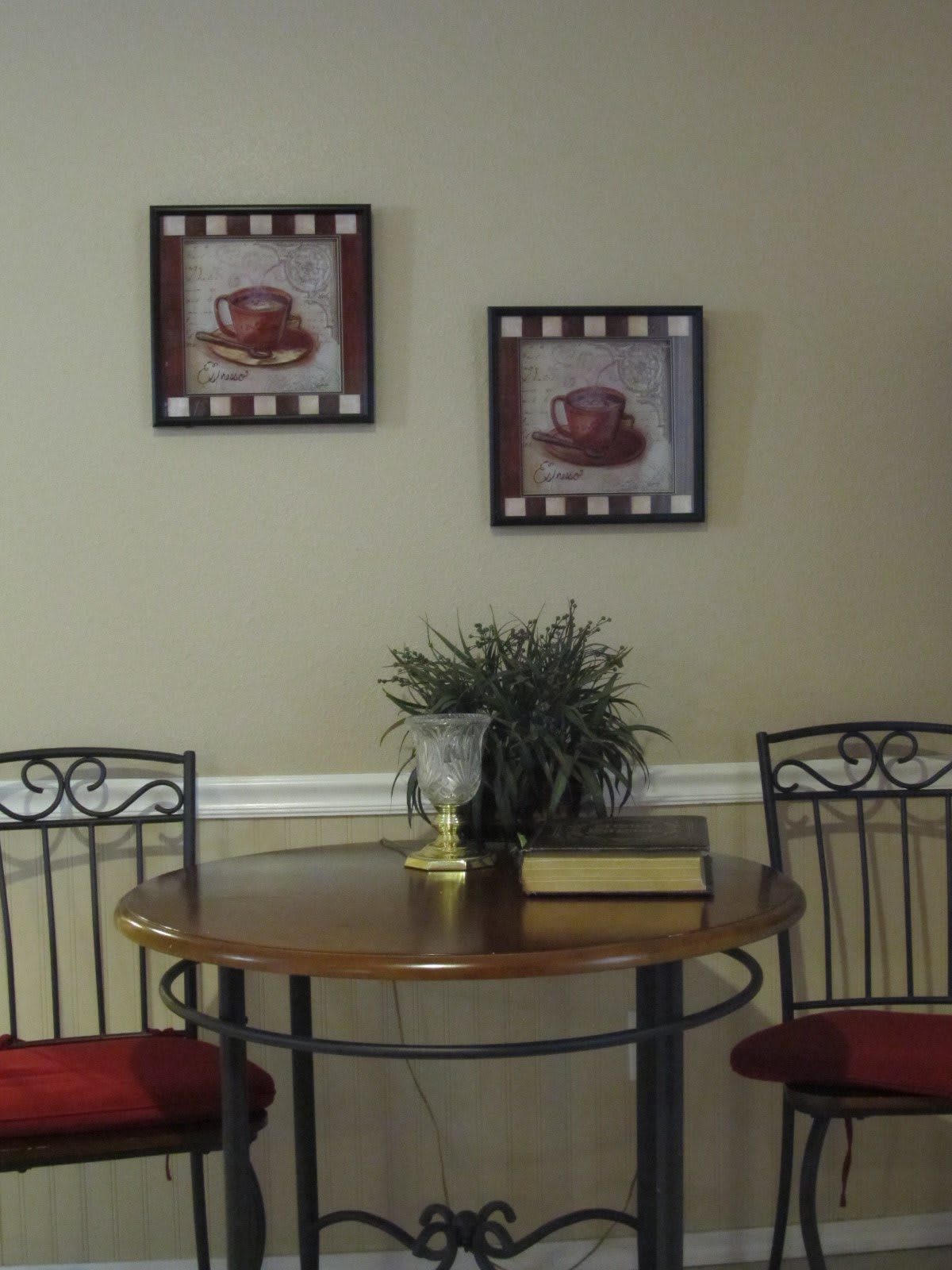 A cozy dining nook with a round wooden table, two chairs, wall art, and a potted plant.