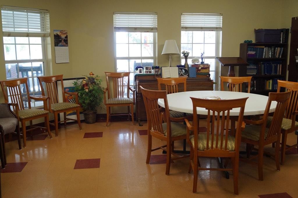 A community meeting room with a round table and wooden chairs, surrounded by windows and various furnishings.