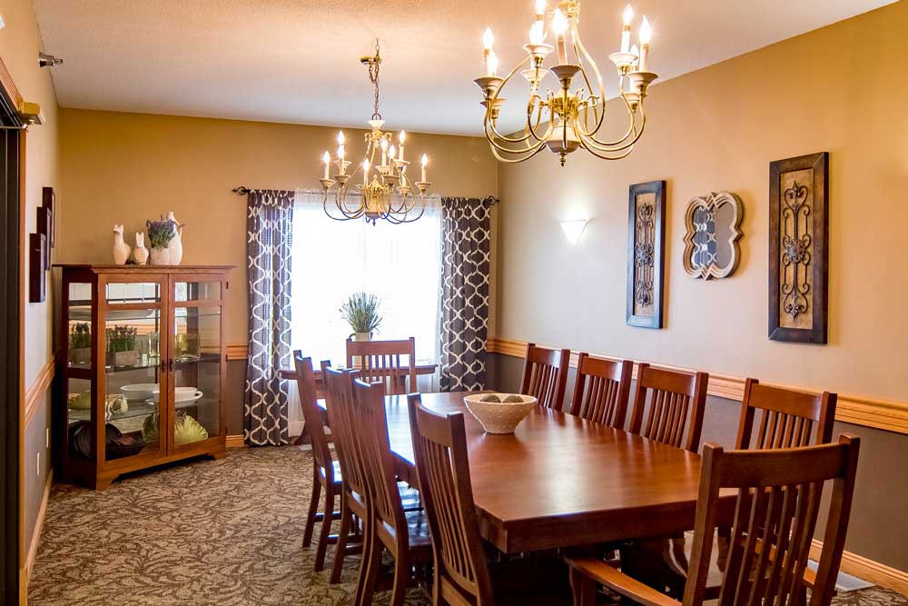 A cozy dining room featuring a wooden table and chairs, illuminated by elegant chandeliers, with decorative elements and natural light filtering through patterned curtains.