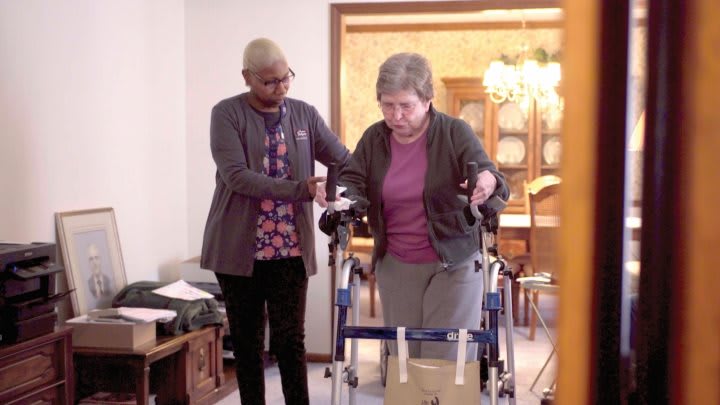 A caregiver assists an elderly woman using a walker in a home setting.
