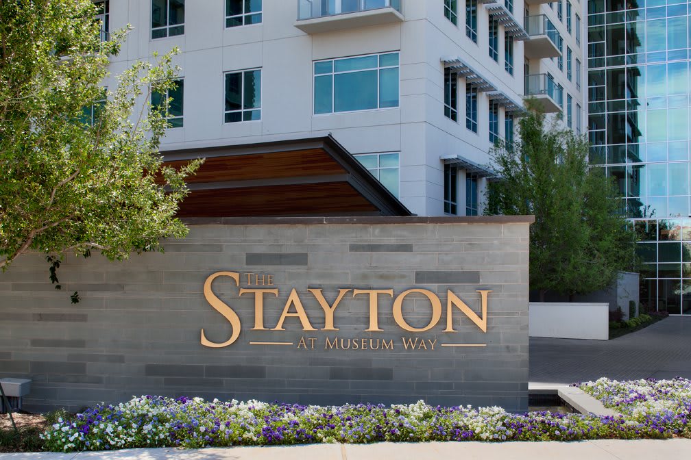 A modern building with a prominent sign for 'The Stayton' surrounded by landscaped greenery.